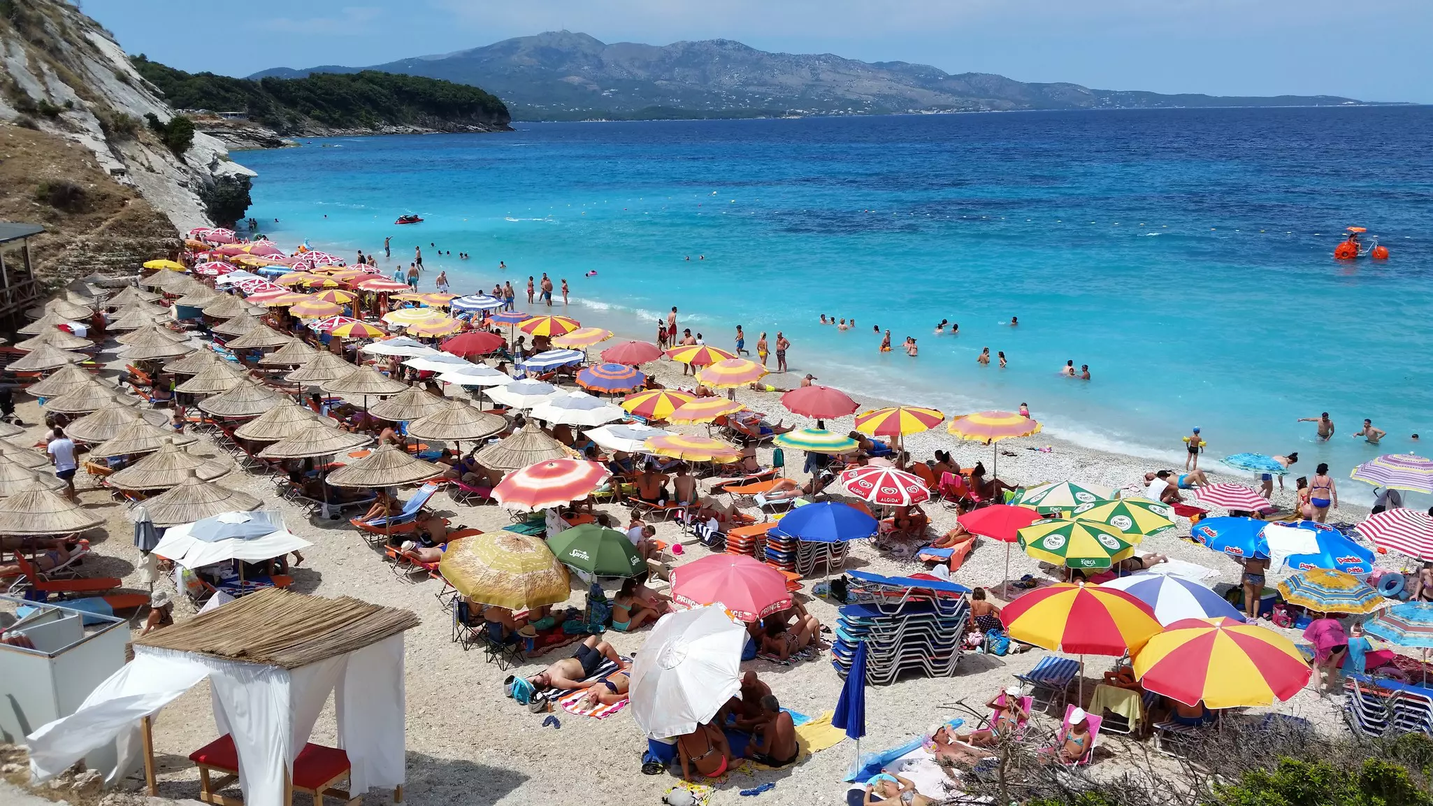 Colorful sun umbrellas are seen from above on a crowded beach facing a blue sea cove.