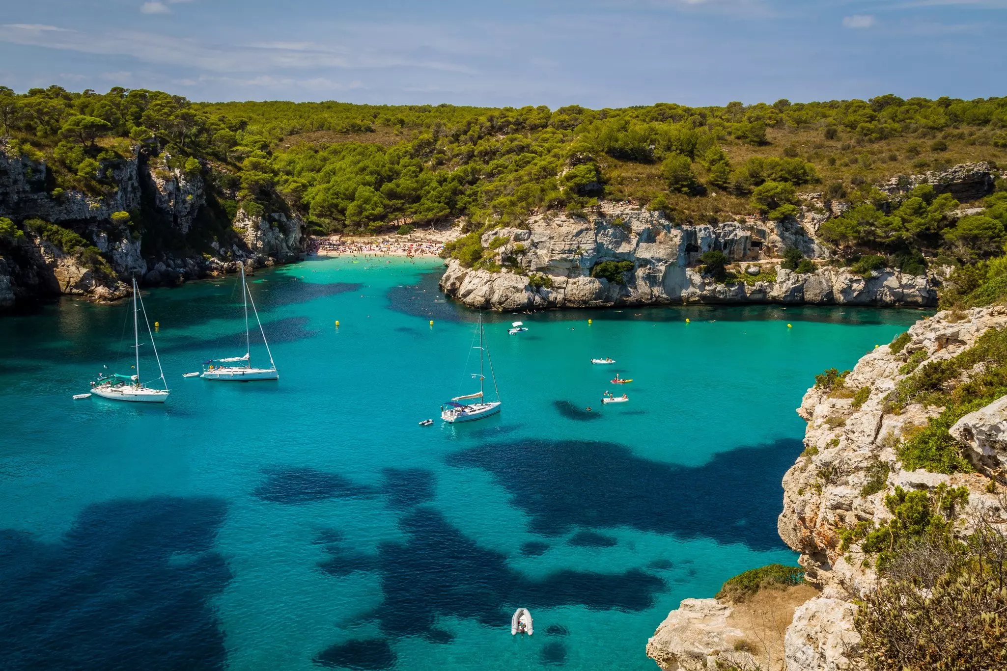 Bay with sailboats in Minorca, Menorca, Spain with a beach in the background crowded with sunseekers