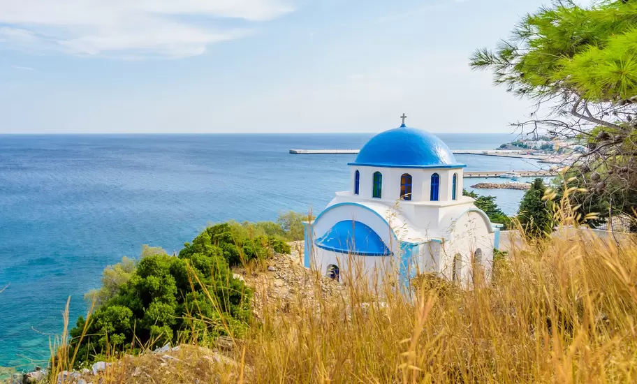 White chapel with blue domes on a dry hillside above the Aegean Sea and a harbour breakwater, Northeastern Aegean Islands