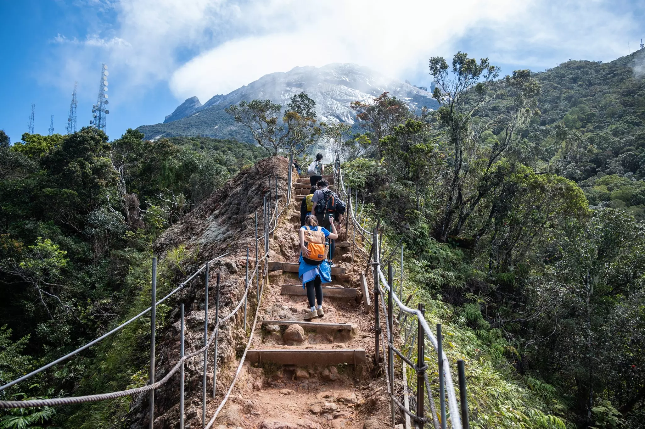 Hikers climb a rocky dirt path toward a mountaintop in Malaysia; stair landings are wedged into the earth, and there is a rope fence on either side of the path.