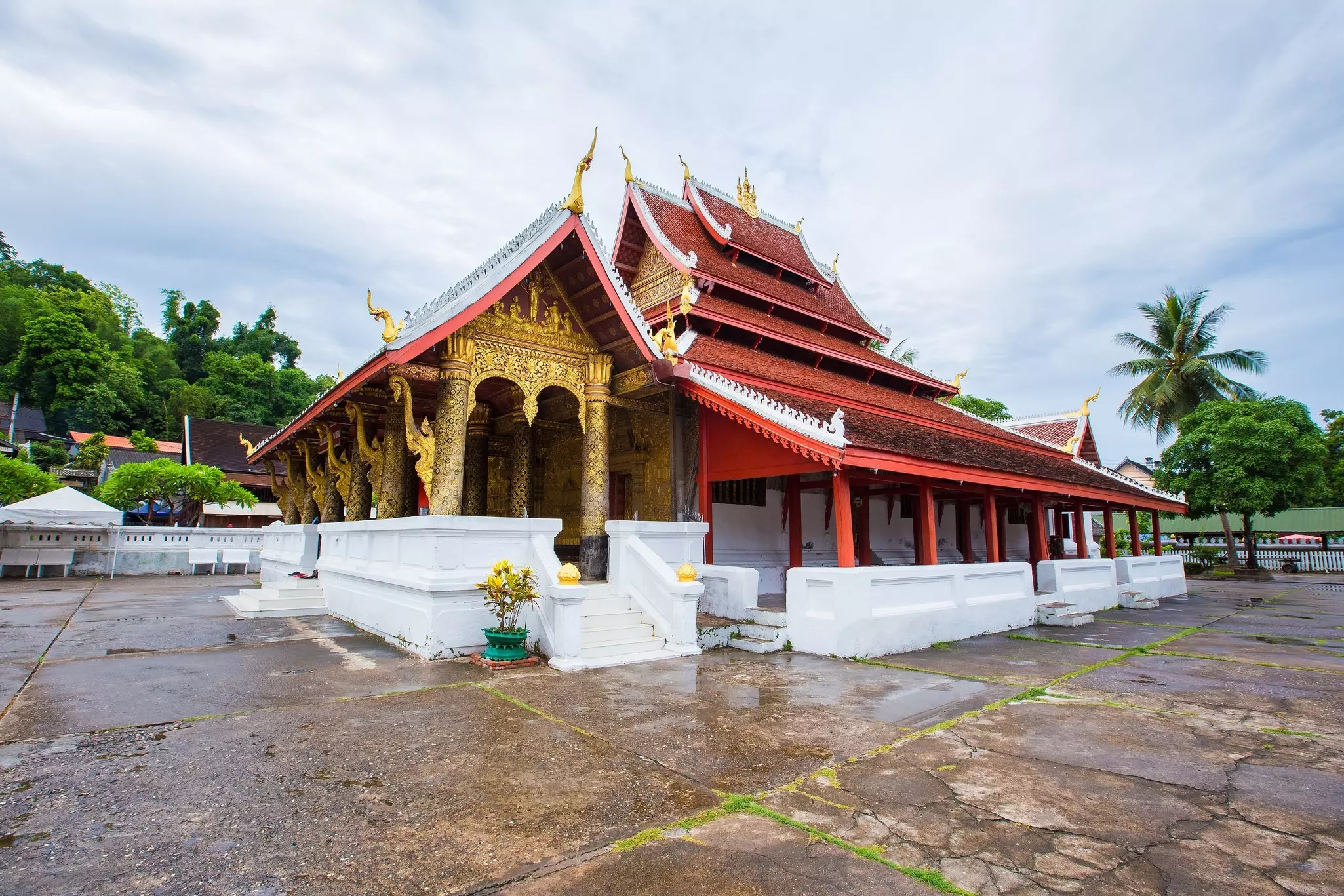 The red, gold and white Wat Mai Suwannaphumaham monastery with a few trees nearby and an overcast sky