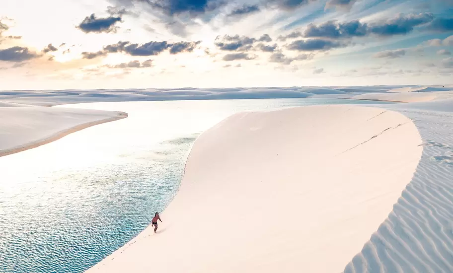 Person climbing a steep, smooth white sand dune beside a pale blue lagoon, with more dunes and a cloudy sky beyond.