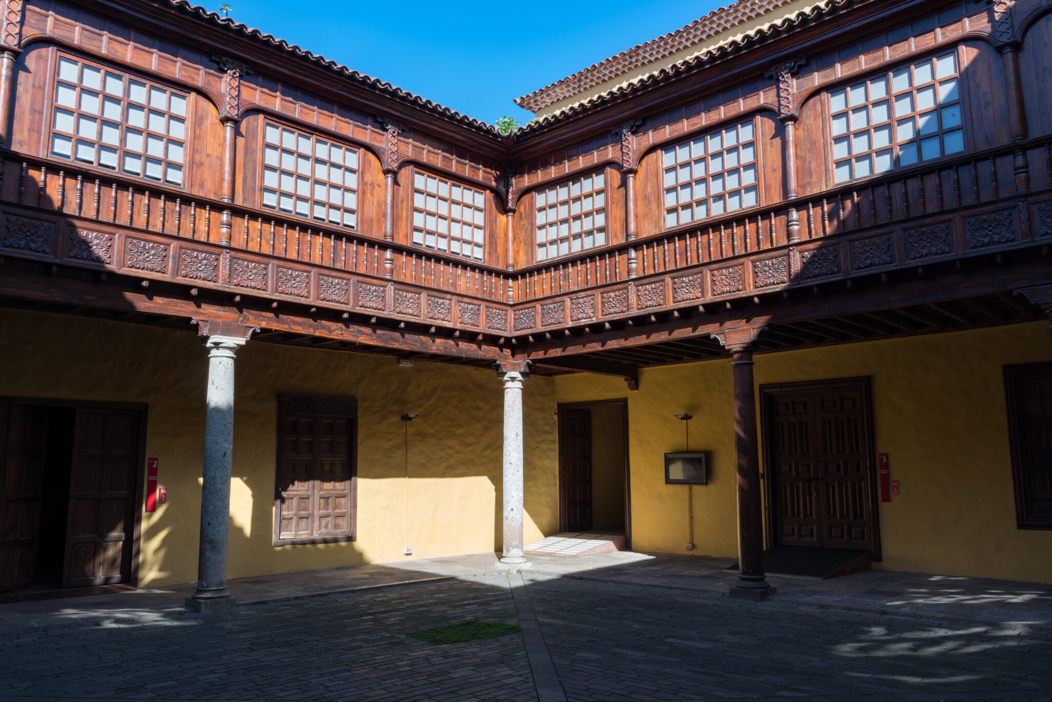 Two-storey courtyard with a carved dark-wood gallery above yellow walls and pale stone columns.
