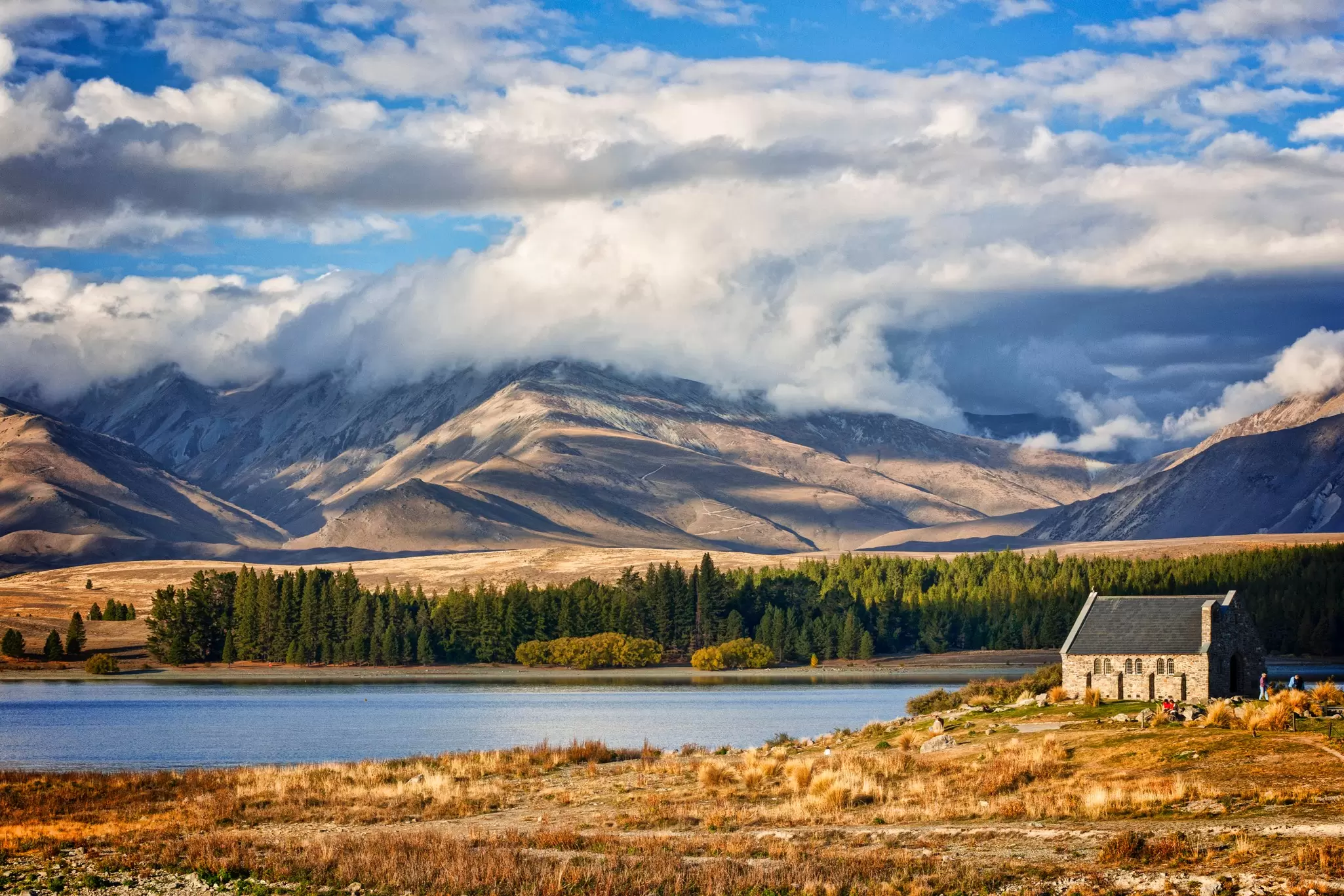 A small stone building on the shore of a lake with towering peaks overlooking them.