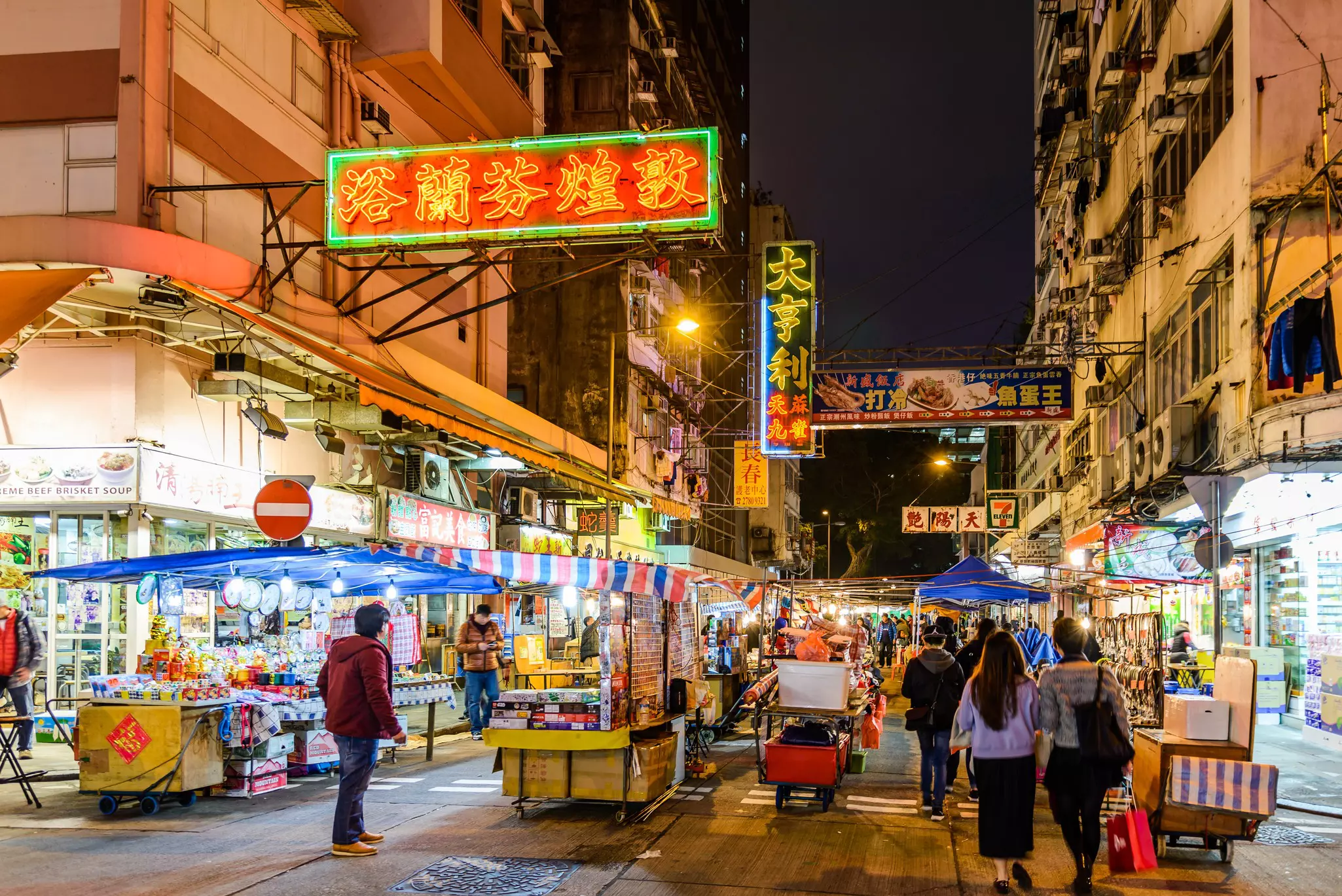 Nighttime view of busy pedestrian alleyway with vendor stalls and markets lining the roadway.