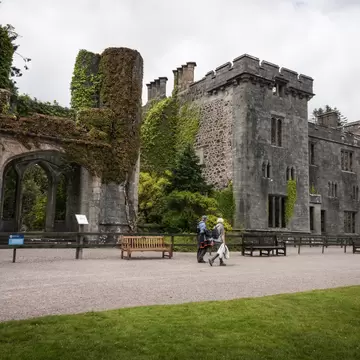 Ivy-covered ruined towers and Gothic arch of Armadale Castle, Skye, with visitors walking a gravel path past benches