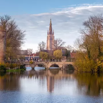 Round brick Powder Tower and arched stone bridge with walkers reflected in Minnewater lake, Church of Our Lady spire behind