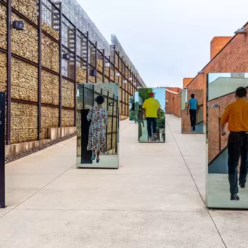 Mirrored pillars printed with life-size photos of walking people line a path between a brick wall and a stone-filled wall
