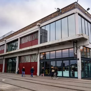 M Shed museum, a former quayside warehouse in Bristol with red doors and 'M shed' banners, blurred walkers on railway tracks