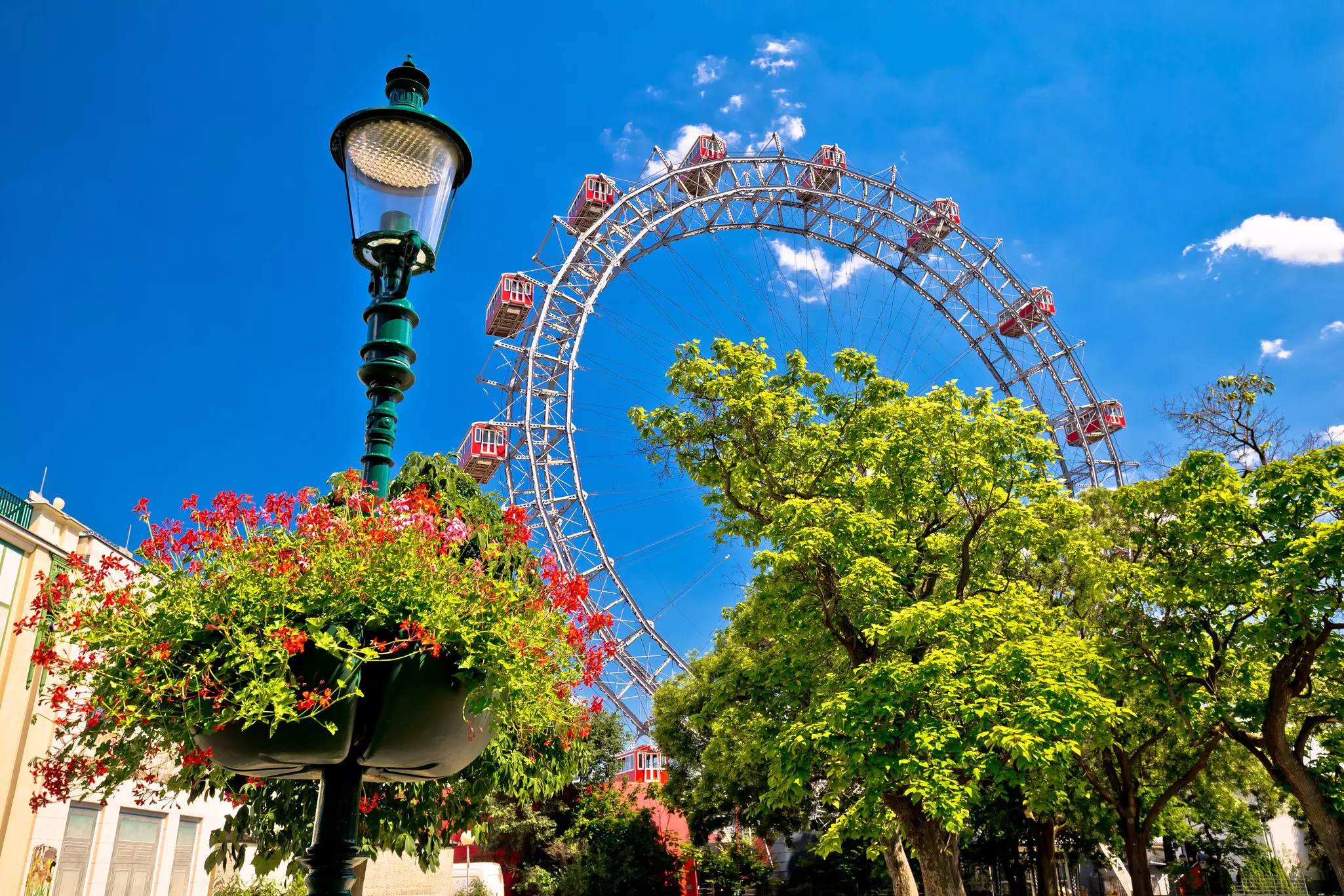 A view of the Riesenrad Ferris wheel at the Prater, with a mostly blue sky, trees, a lamp post and some buildings.