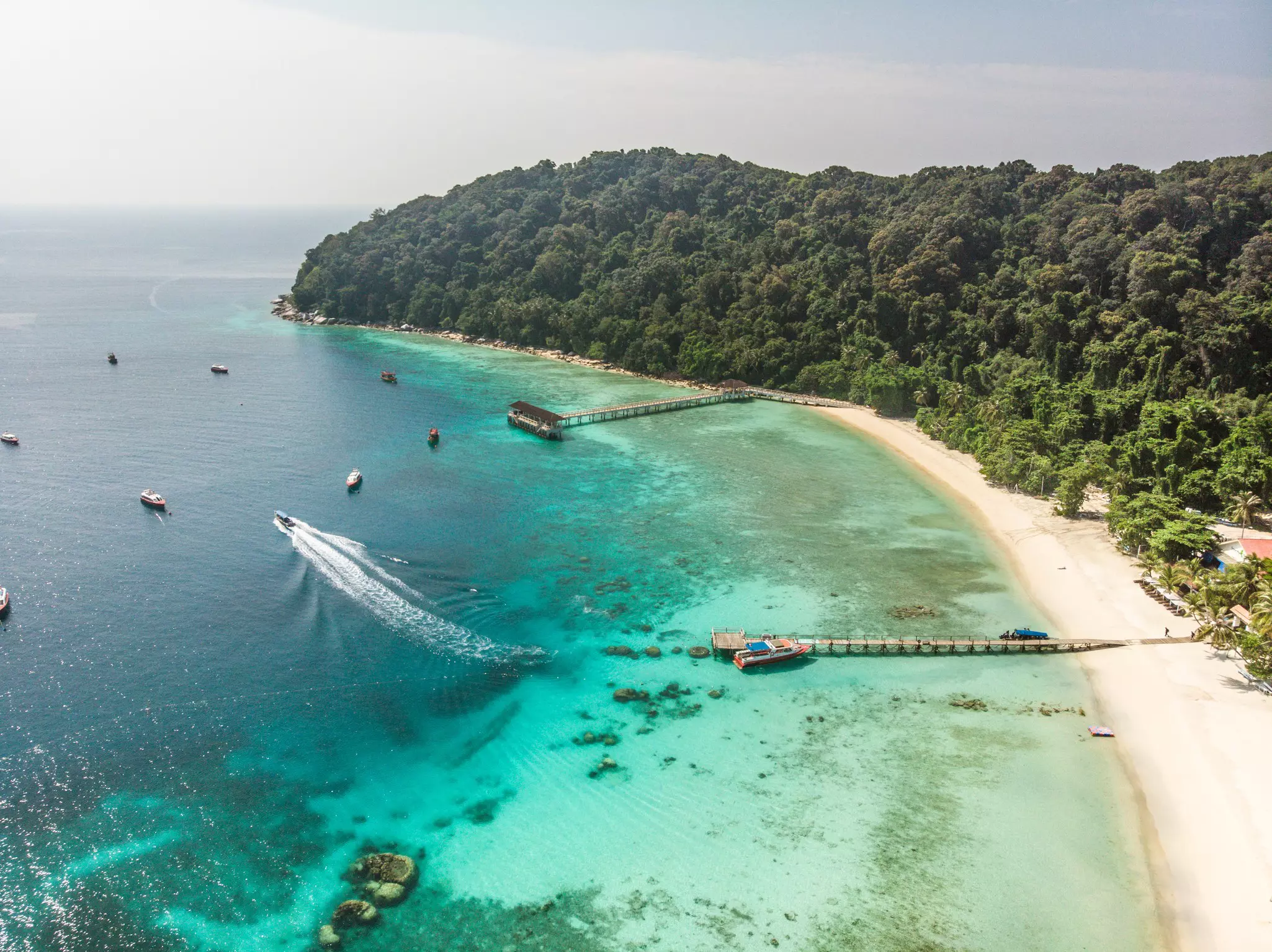 An aerial soot of boats in the clear blue waters off of Pulau Lang Tengah Island, with docks off a white-sand beach and a slope covered in green palm trees, Malaysia