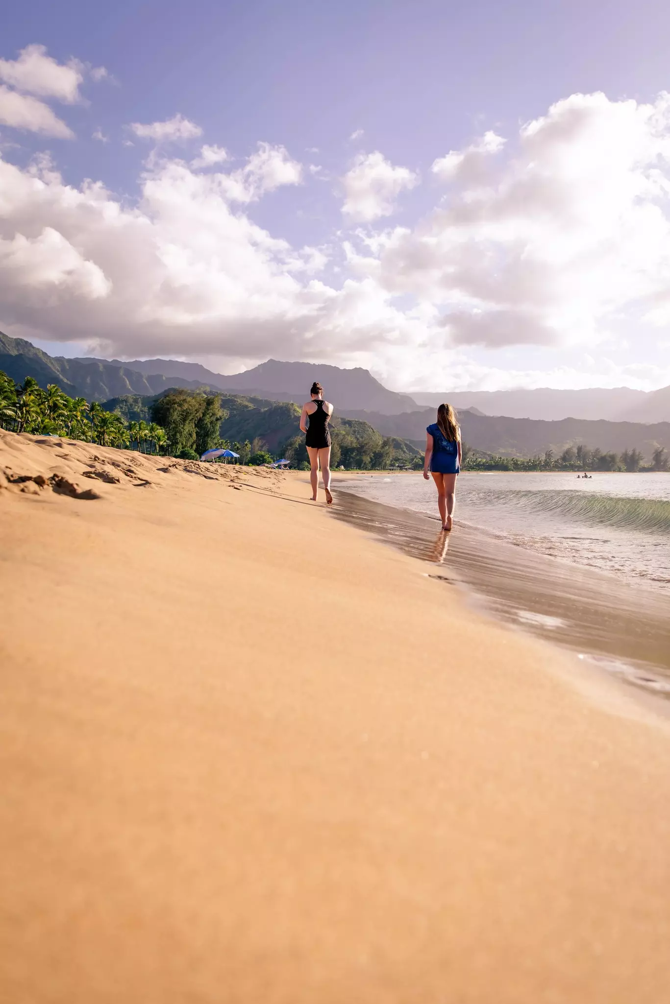 Two walking or strolling along the shore of a white sand beach with green tree covered hills in background. Two colorful beach umbrellas are in the distance.