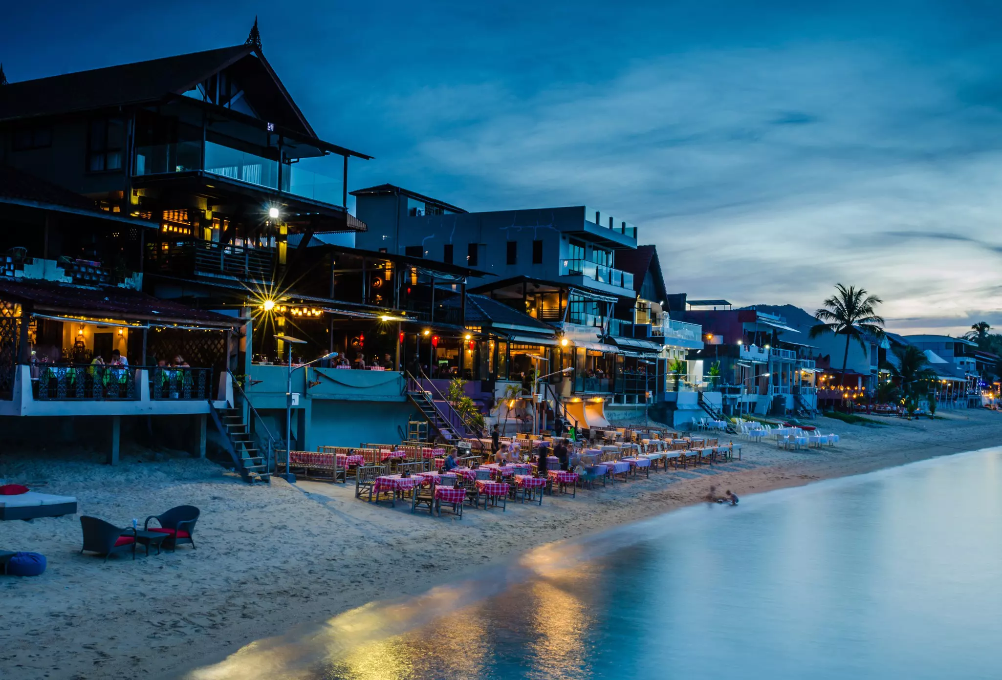 Tables set out on the beach backed by restaurants as twilight falls, lights come on and diners come out for dinner.