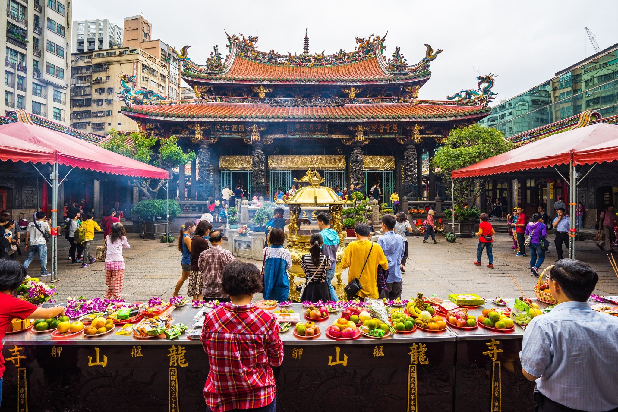 People milling about Longshan Temple in Taiwan with a table in front with offerings of fresh fruit and other food.