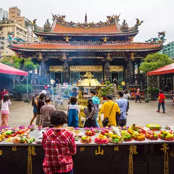 People milling about Longshan Temple in Taiwan with a table in front with offerings of fresh fruit and other food.