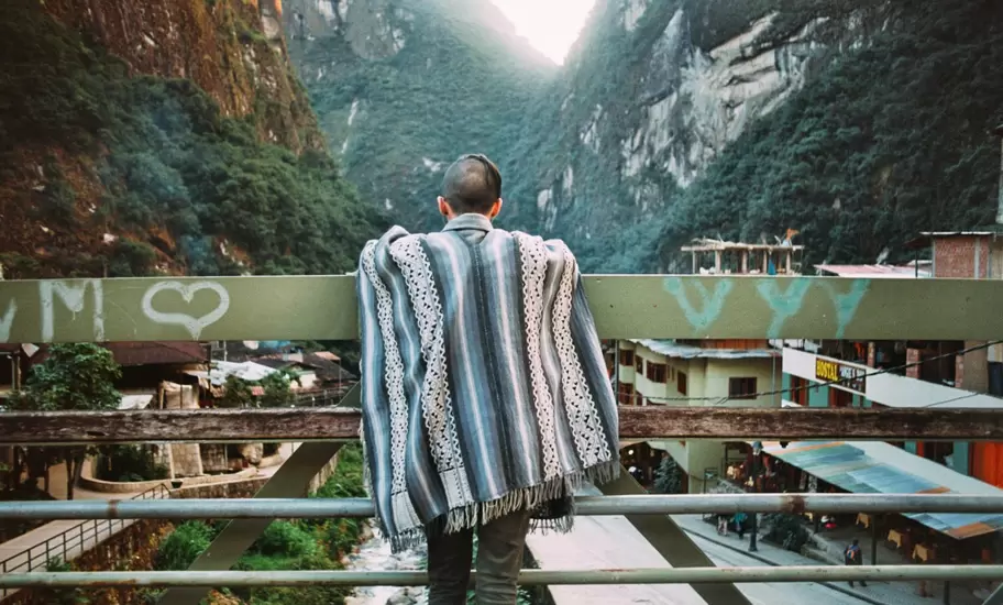A man wearing a Peruvian poncho leaning on the rails of a bridge at Aguas Calientes.