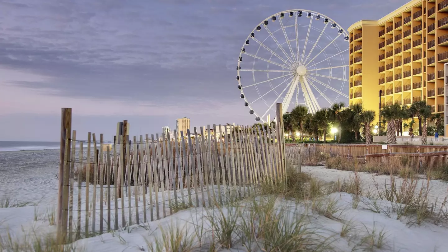 A fence in the dunes along the Boardwalk of Myrtle Beach at dusk, with the SkyWheel and apartment buildings in the distance