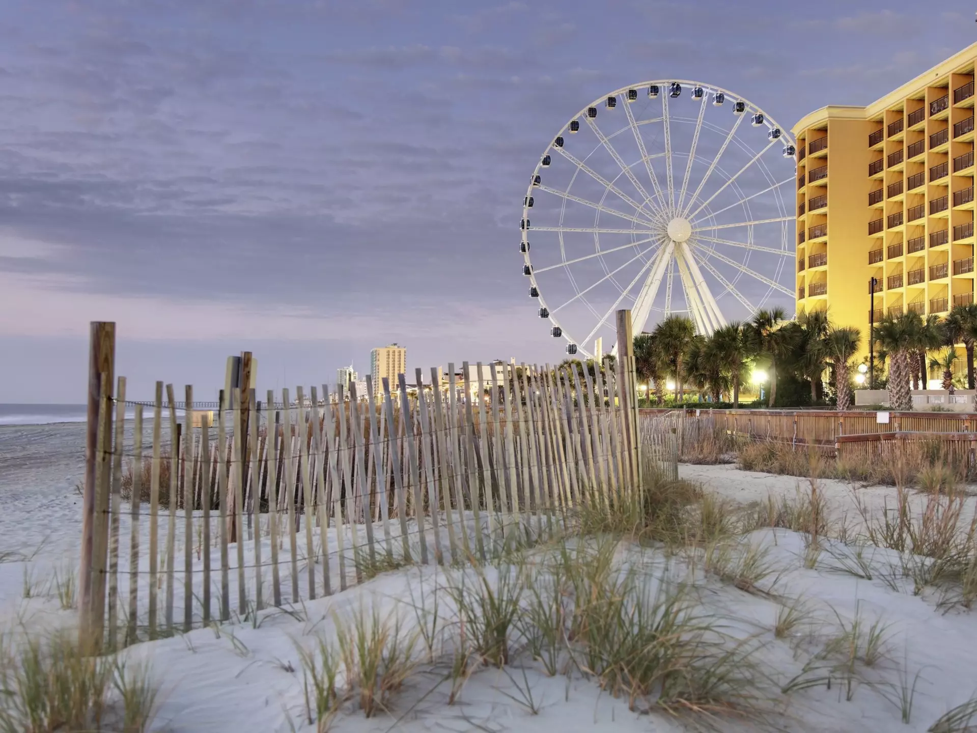 A fence in the dunes along the Boardwalk of Myrtle Beach at dusk, with the SkyWheel and apartment buildings in the distance