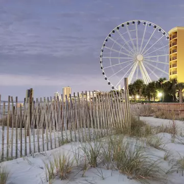 A fence in the dunes along the Boardwalk of Myrtle Beach at dusk, with the SkyWheel and apartment buildings in the distance