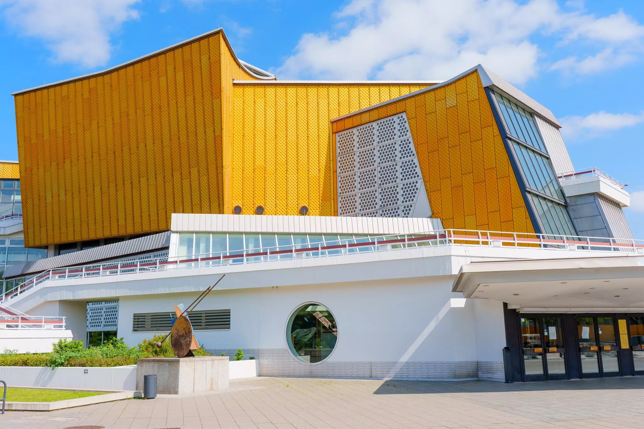 A large, modern building, white at the bottom and yellow at top, on a stone block plaza on a sunny day.