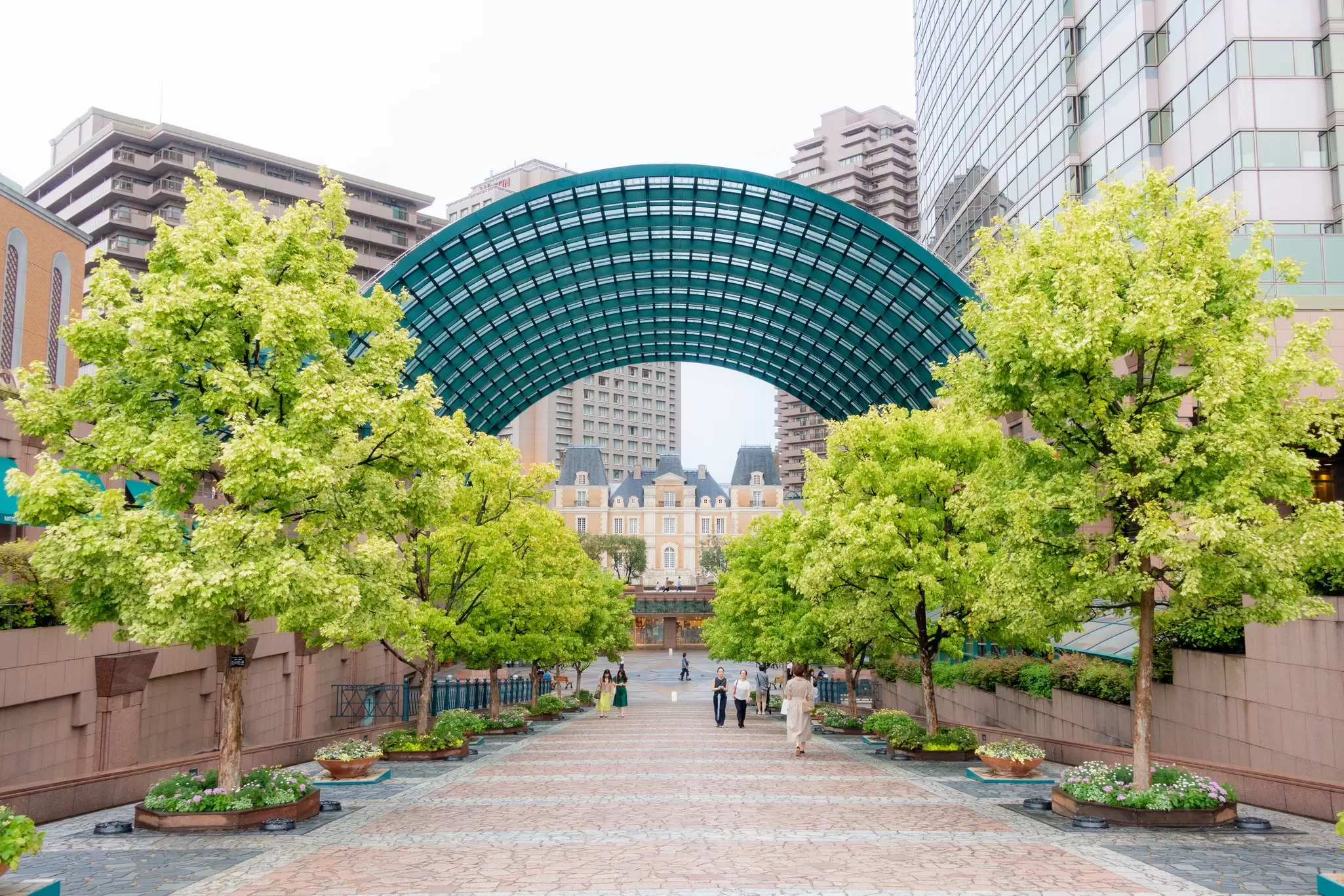 People walking on a plaza lined with green trees and a green archway over it