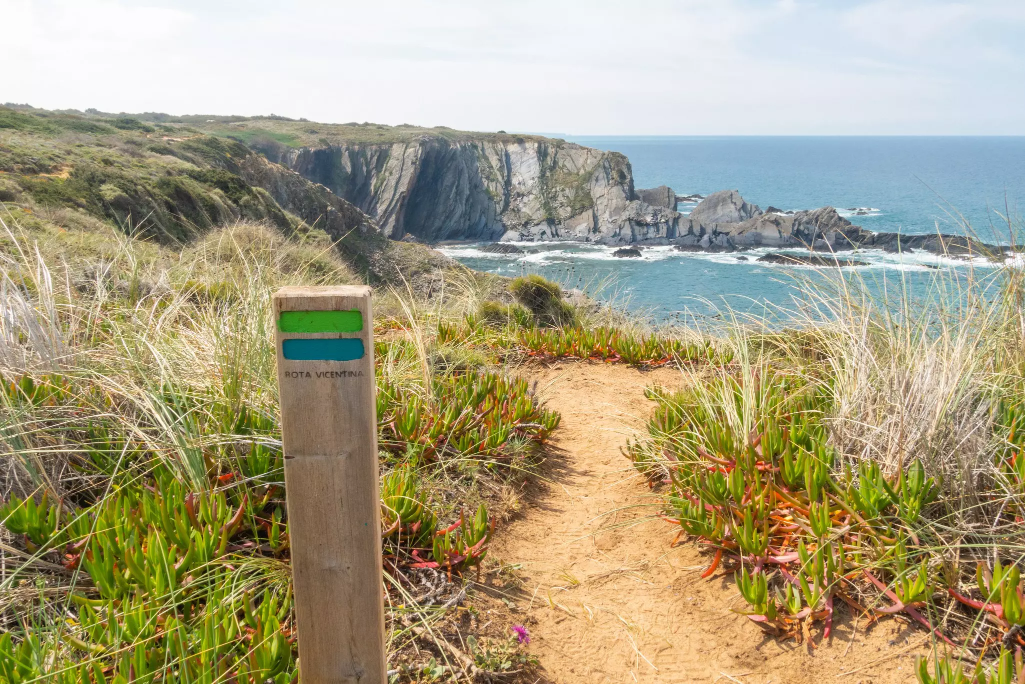 A wooden post with a green and blue line painted on it marks the way down a sandy coastal path on a cliff-top hiking route.