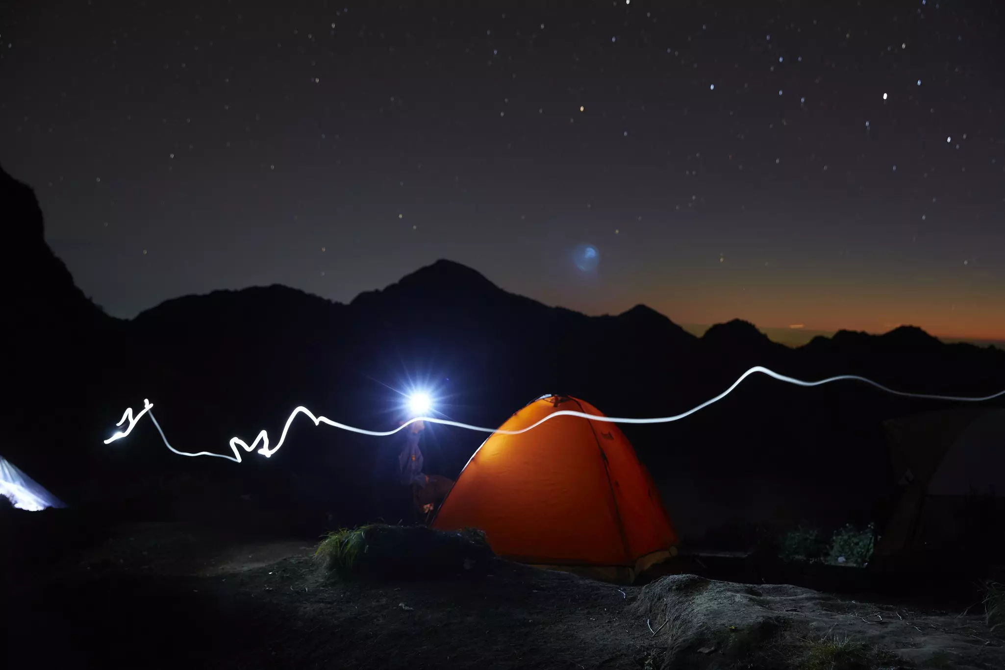 An orange tent shines brightly in the dark night with a mountain in the background