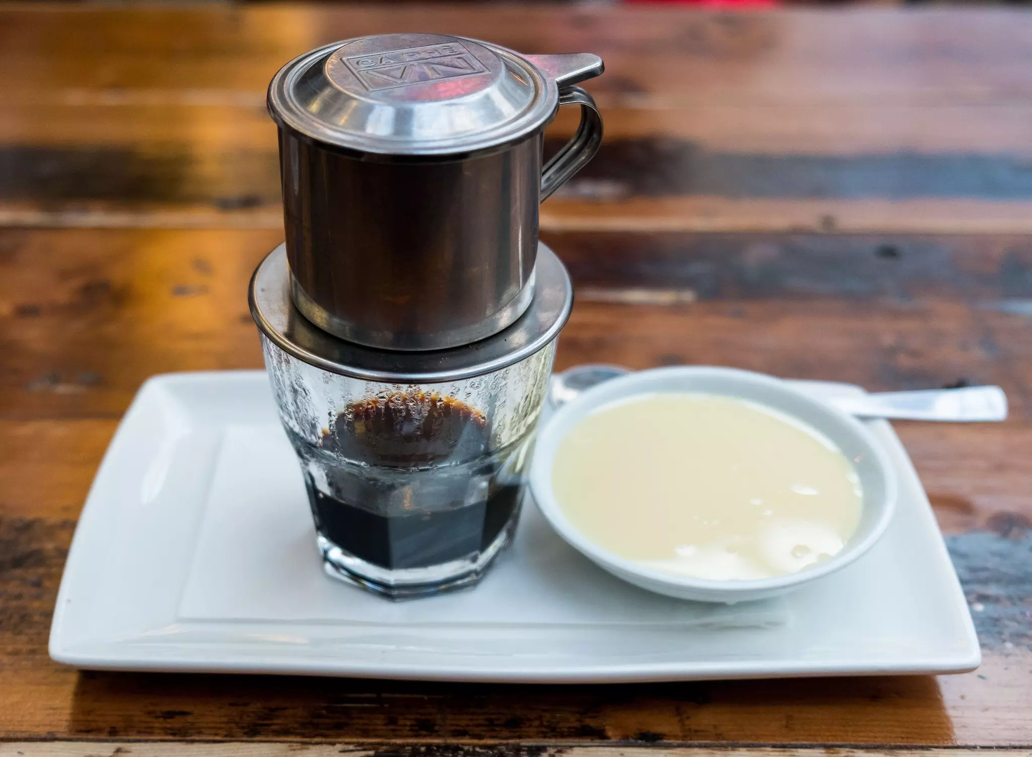 A cup of Vietnamese coffee with a saucer of cream on a larger platter resting on a wooden table.