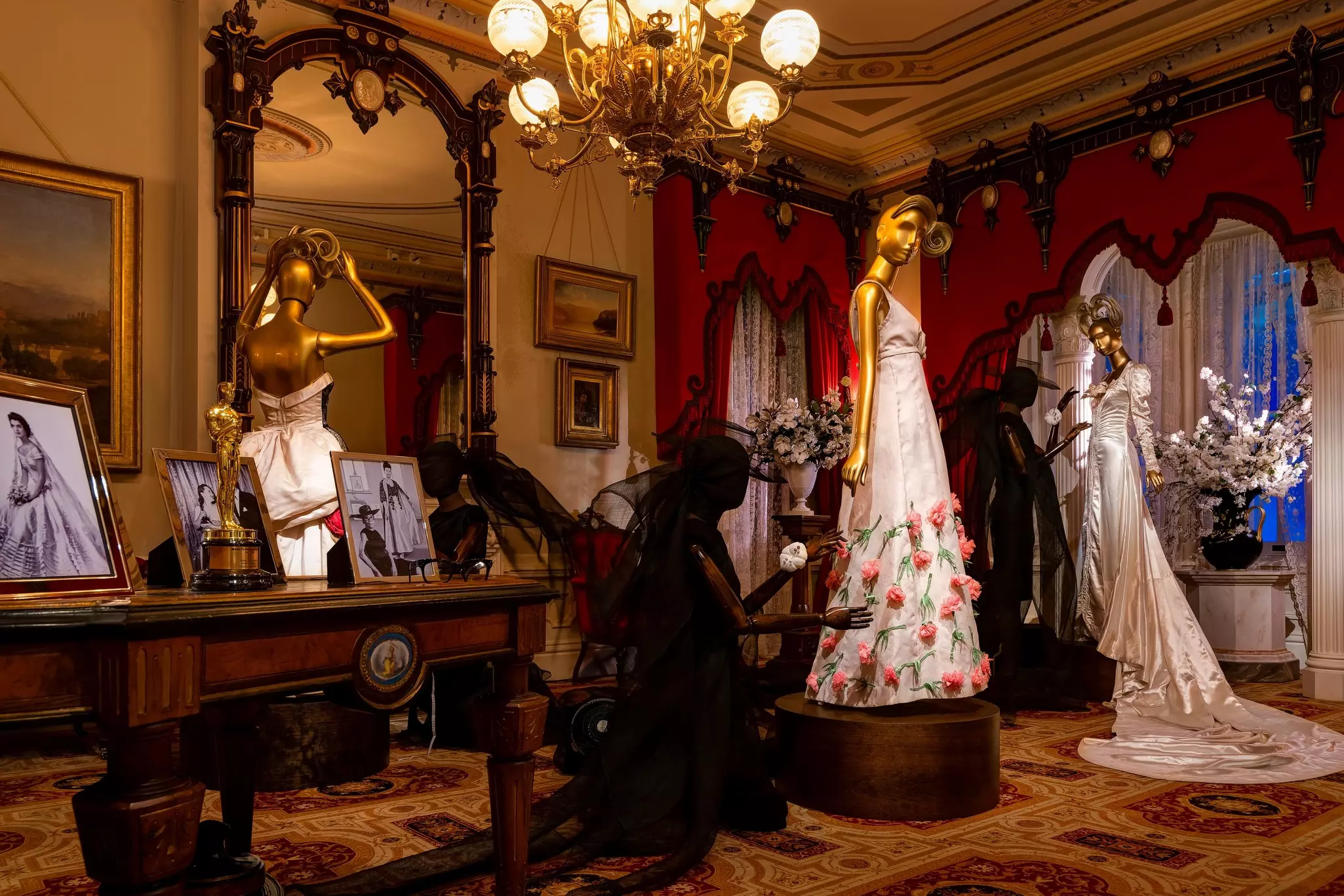 Gold mannequins wearing formal gowns in a room with windows covered in deep red drapes, and a large mirror with a thick gold frame