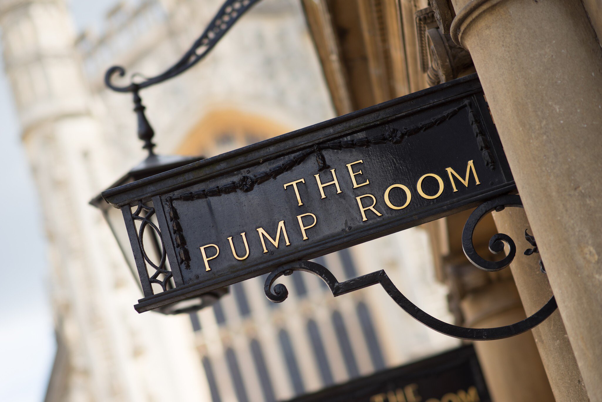 Black iron hanging sign reading 'The Pump Room' in gold letters beside a lantern, with Bath Abbey blurred behind