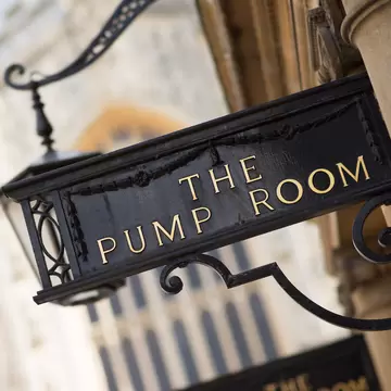 Black iron hanging sign reading 'The Pump Room' in gold letters beside a lantern, with Bath Abbey blurred behind