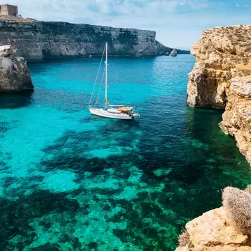 White sailboat on turquoise sea in a rocky cove, pale limestone cliffs on both sides and a stone tower on the far clifftop.