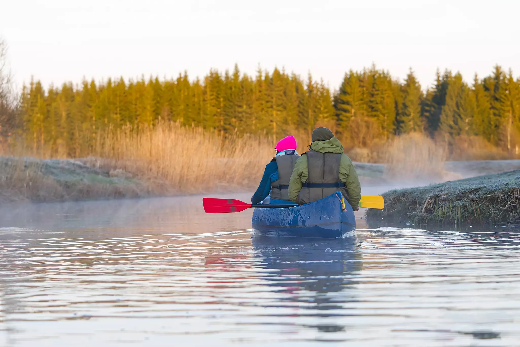 Two people in a kayak paddle their way along a river towards some woodland on a chilly morning.