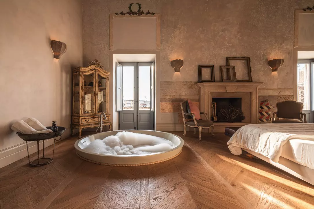 A large bedroom in a historic building with original features including a stylish wooden floor, balcony doors and stone fireplace. A sunken bathtub sits adjacent to the large bed.