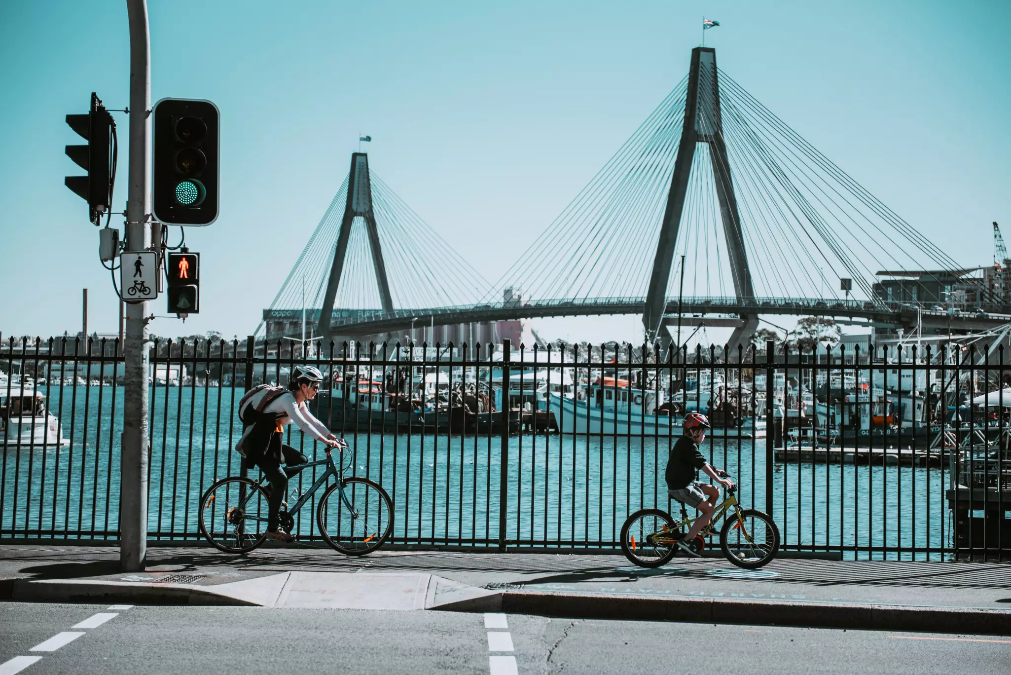 A woman and a child ride bikes on the sidewalk near a harbor. A large suspension bridge passes behind them.