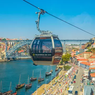Cable car cabin over a crowded riverside street in Vila Nova de Gaia, wooden boats on the Douro and an iron arch bridge.