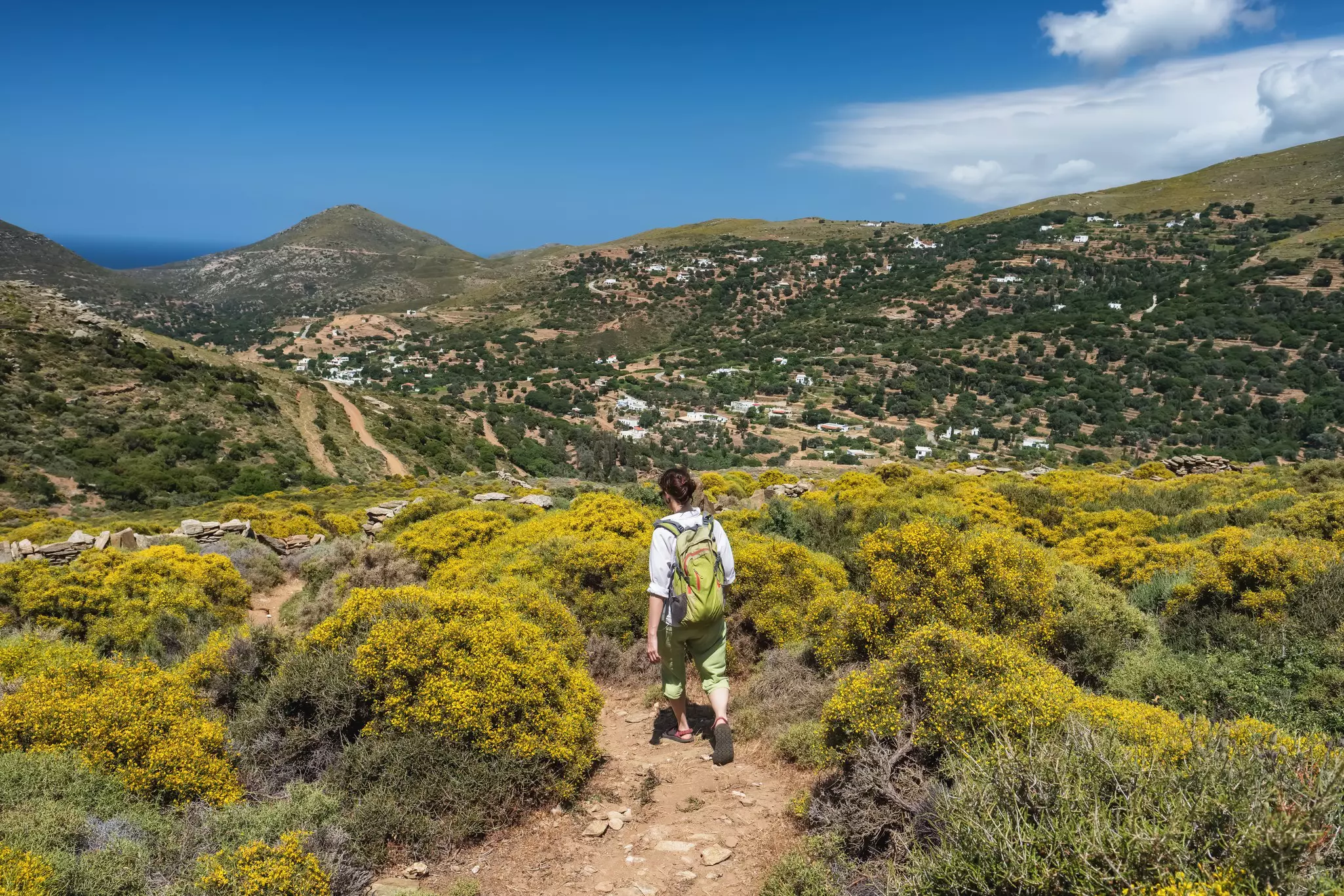 A hiker walking on a dirt path through green scrub; there are scattered houses on the hillside and a bright blue sky.