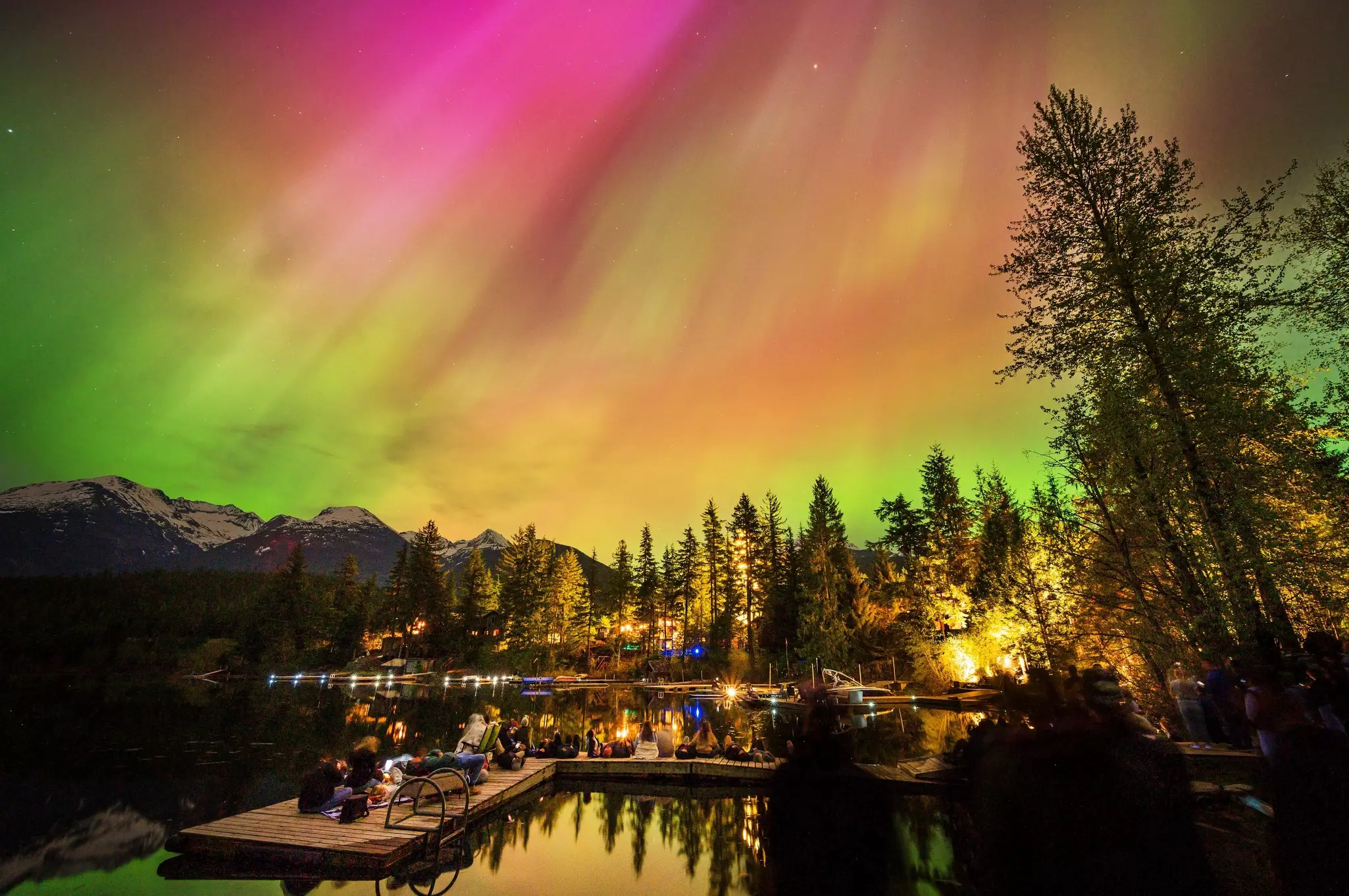 Crowds of people on a dock in the woods watch the pink, yellow, and green Northern Lights shine brightly above.