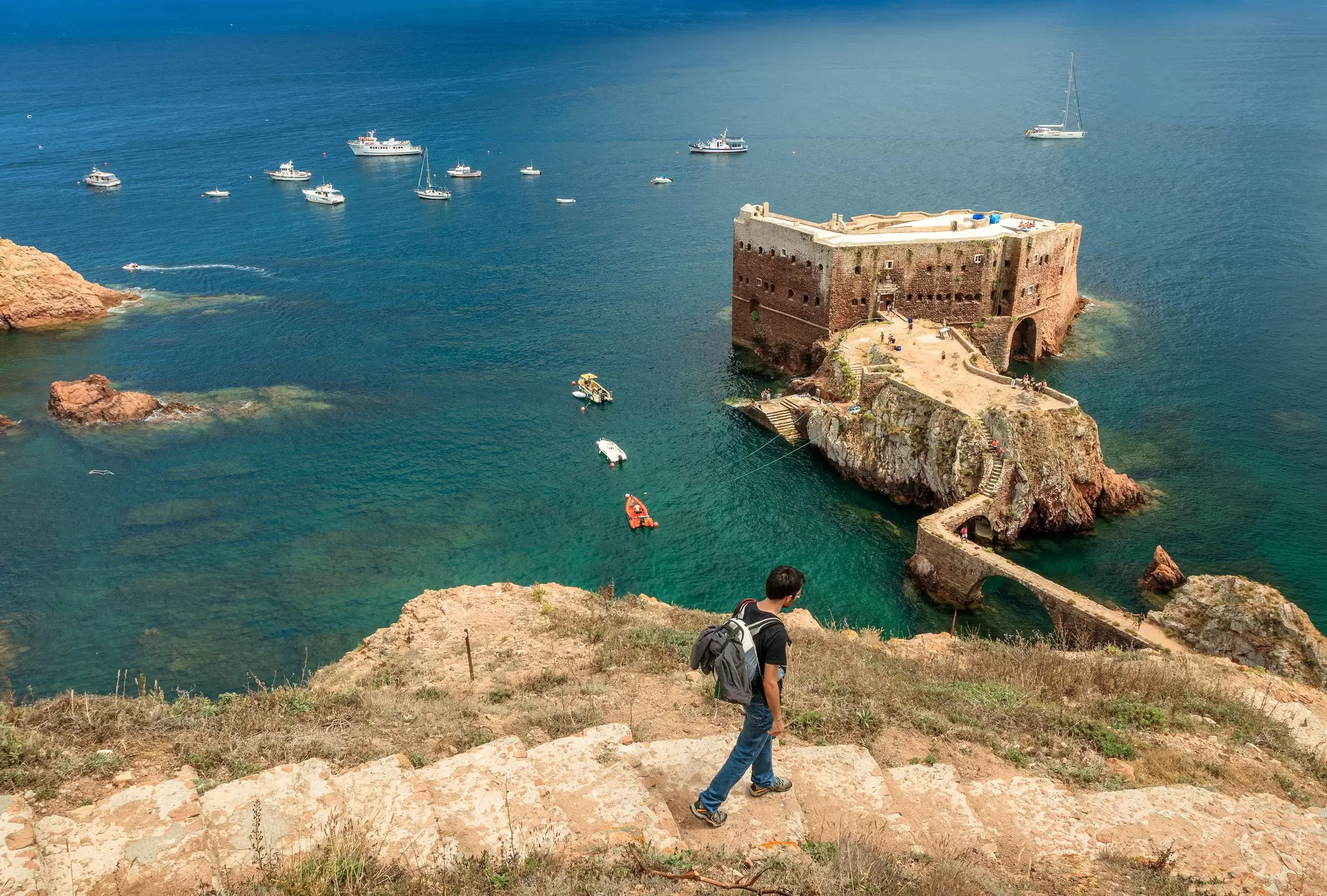 Berlenga island, Portugal - July 09, 2017: Young man with backpack walking down the access path to Fort St. John the Baptist of Berlengas, with the fort, sea and boats in the background.  License Type: media  Download Time: 2023-08-15T10:31:47.000Z  User: clairenaylor  Is Editorial: Yes  purchase_order:   