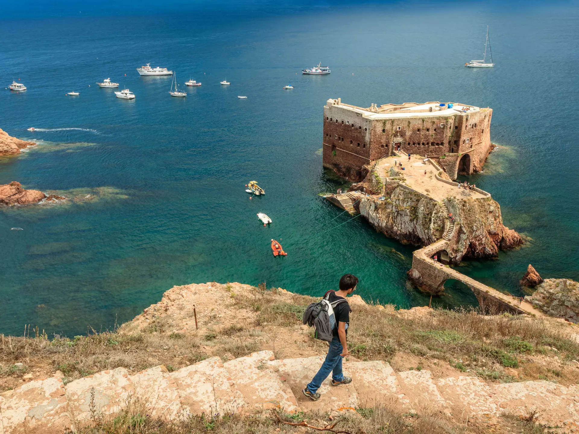 Berlenga island, Portugal - July 09, 2017: Young man with backpack walking down the access path to Fort St. John the Baptist of Berlengas, with the fort, sea and boats in the background.  License Type: media  Download Time: 2023-08-15T10:31:47.000Z  User: clairenaylor  Is Editorial: Yes  purchase_order:   