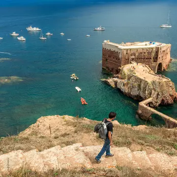 Berlenga island, Portugal - July 09, 2017: Young man with backpack walking down the access path to Fort St. John the Baptist of Berlengas, with the fort, sea and boats in the background.  License Type: media  Download Time: 2023-08-15T10:31:47.000Z  User: clairenaylor  Is Editorial: Yes  purchase_order:   