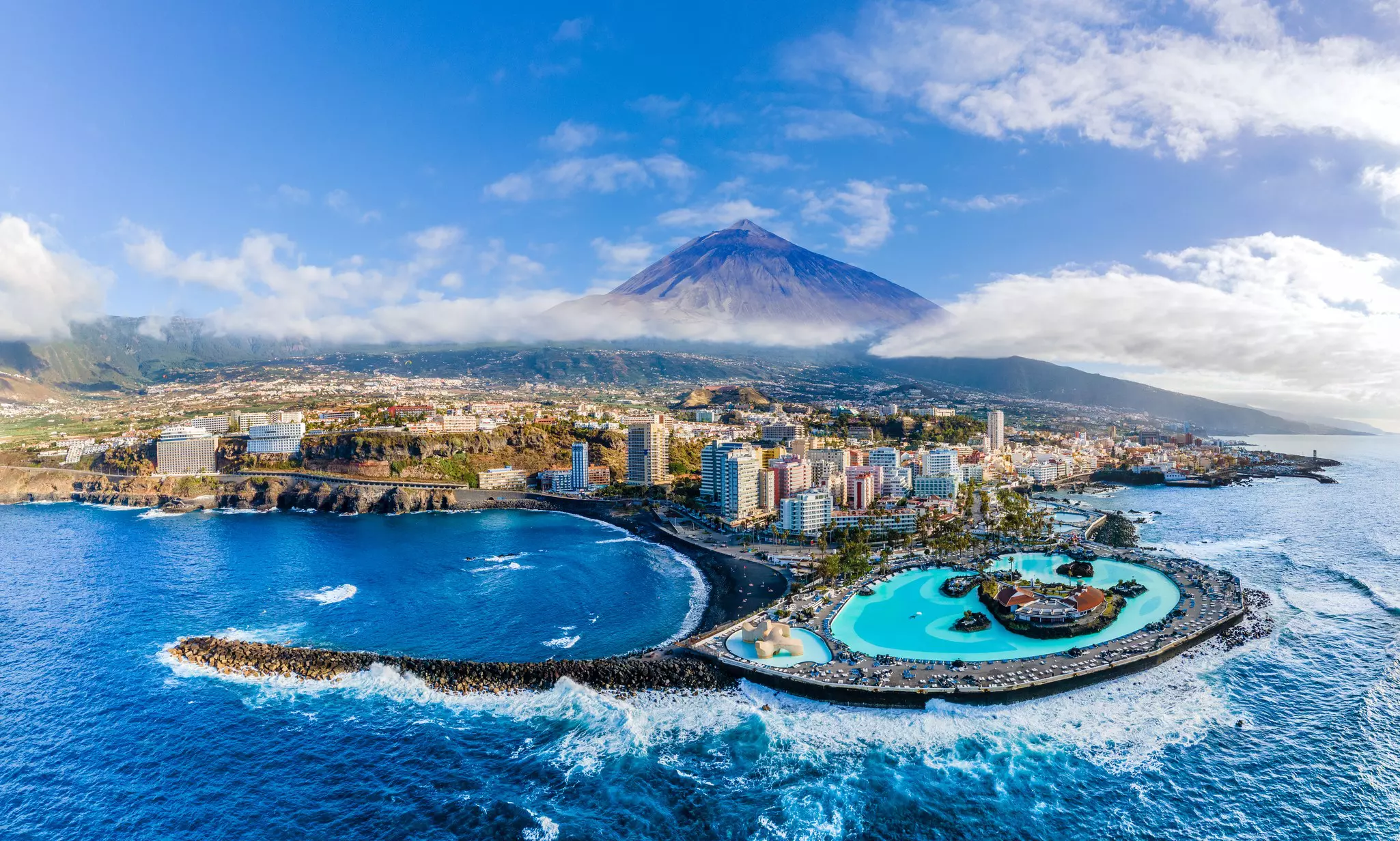 A pointed volcano rises above a layer of mist. Below is a resort with pools and surf crashing on the rugged shores