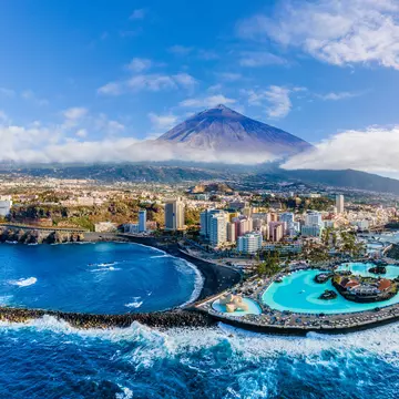 A pointed volcano rises above a layer of mist. Below is a resort with pools and surf crashing on the rugged shores