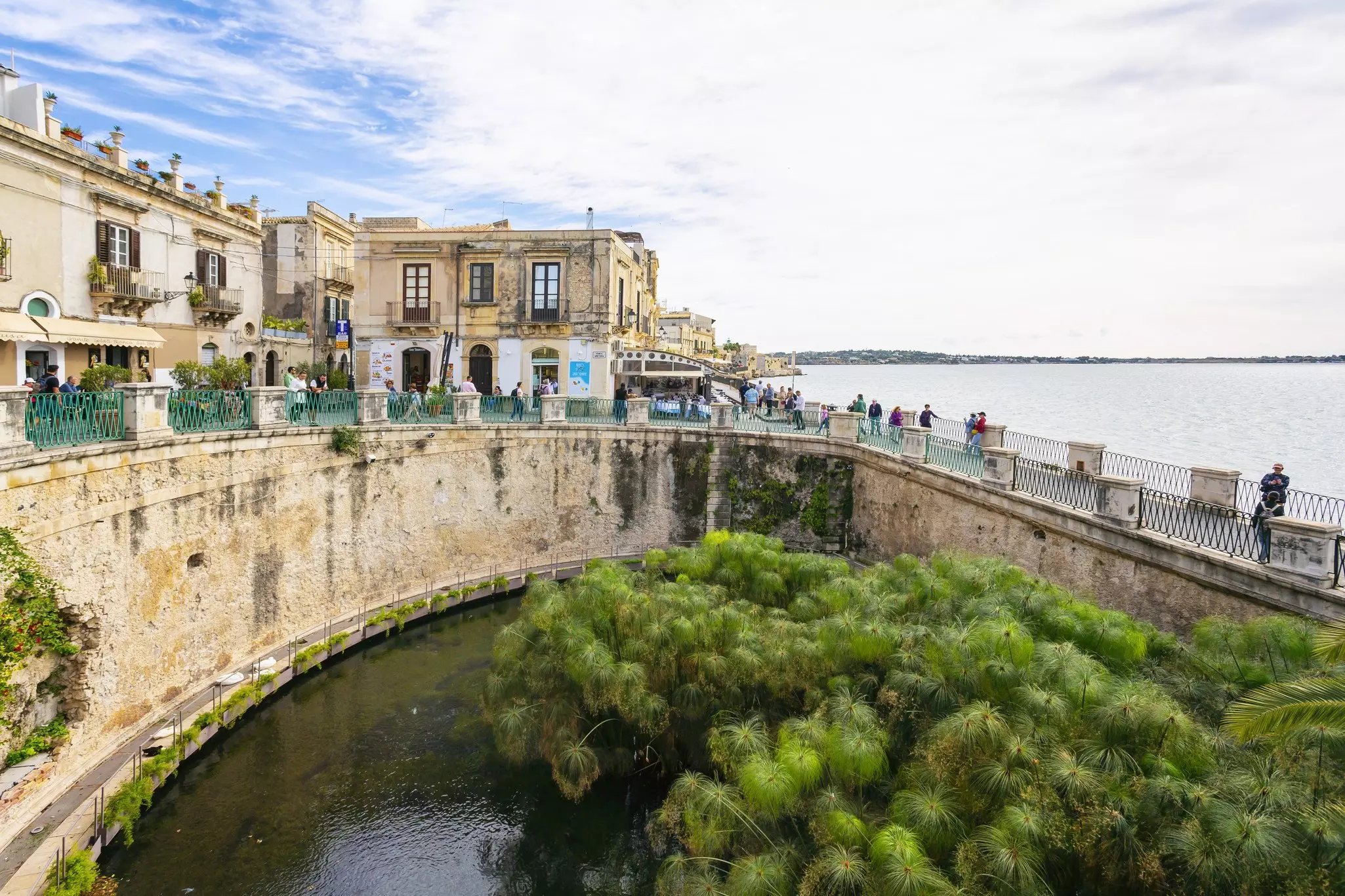 Water bound by a curved stone wall with buildings to the left and the ocean to the right on a partly cloudy day.