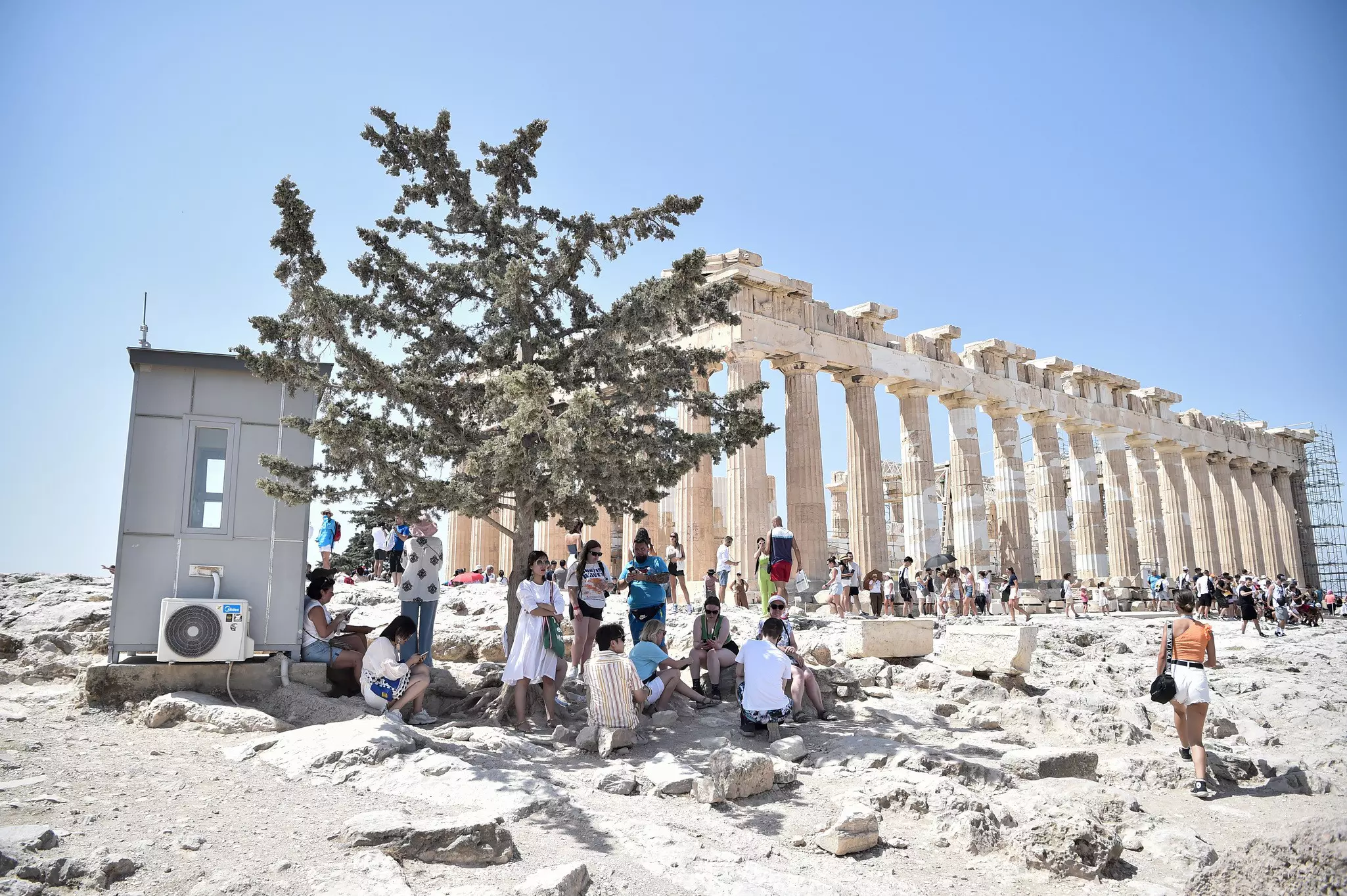 Atop the Acropolis ancient hill with Parthenon temple in background, tourists shelter from the sun under a tree