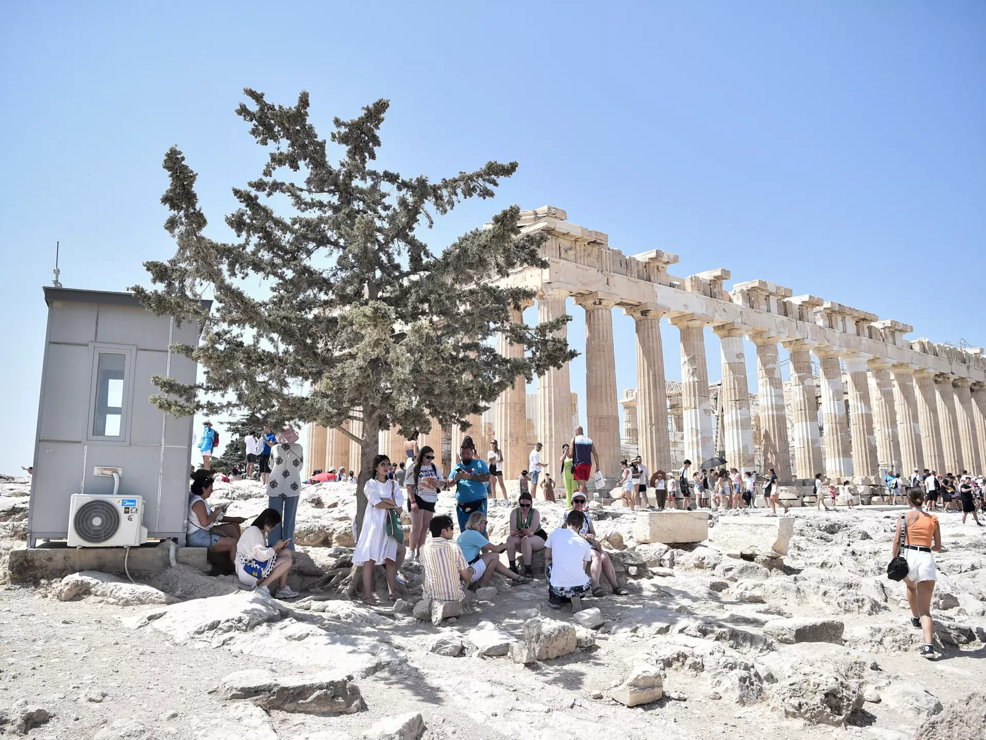 Atop the Acropolis ancient hill with Parthenon temple in background, tourists shelter from the sun under a tree