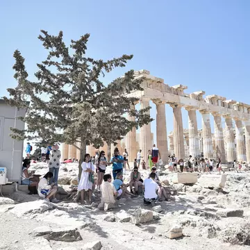 Atop the Acropolis ancient hill with Parthenon temple in background, tourists shelter from the sun under a tree
