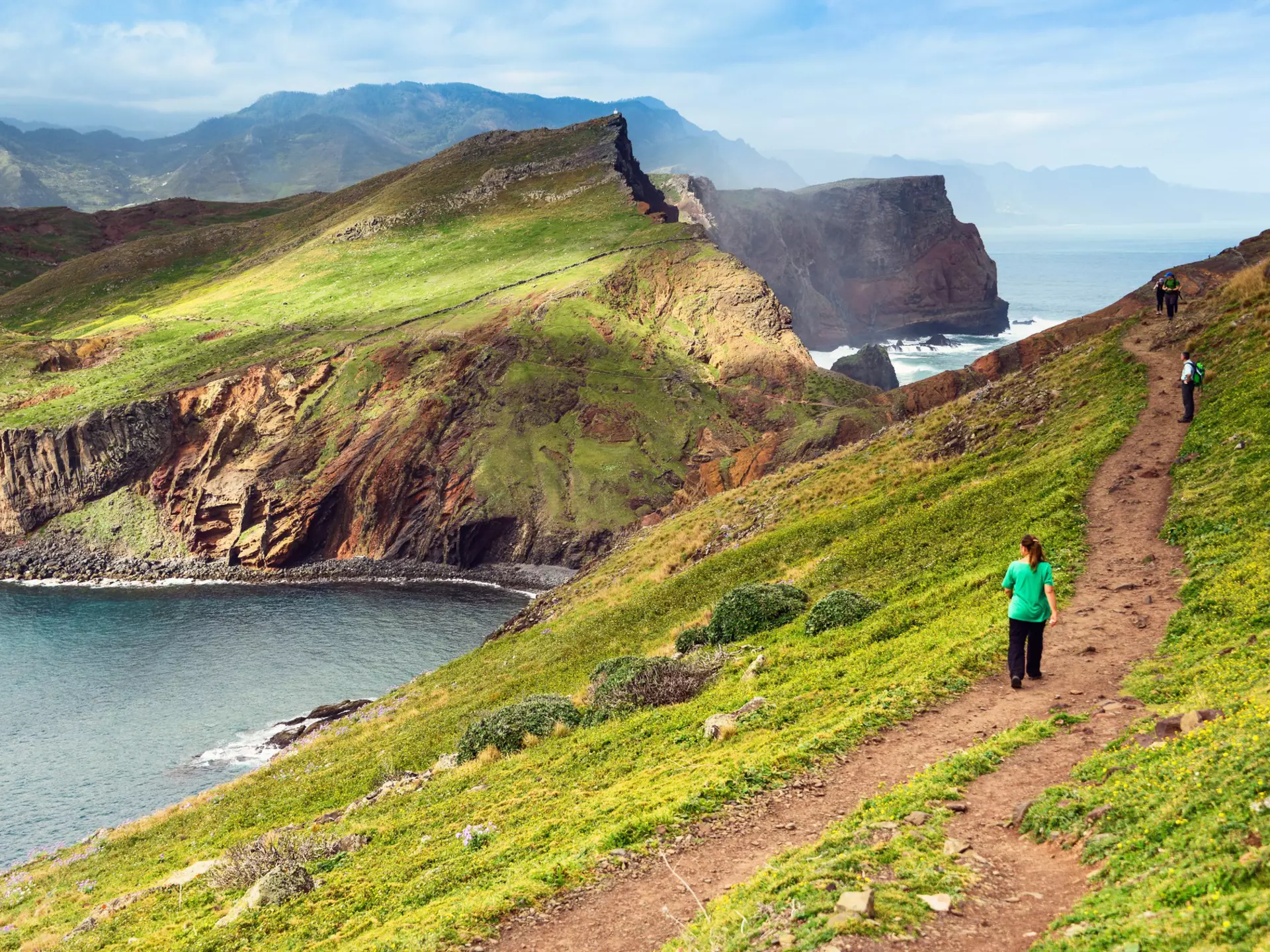 A few hikers spaced well apart walk on a dirt path on a grassy hill by the coast; one is looking down toward the sea, and there are rugged mountains in the distance.