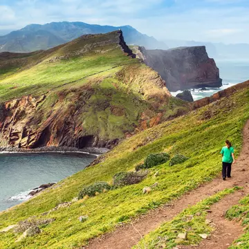 A few hikers spaced well apart walk on a dirt path on a grassy hill by the coast; one is looking down toward the sea, and there are rugged mountains in the distance.