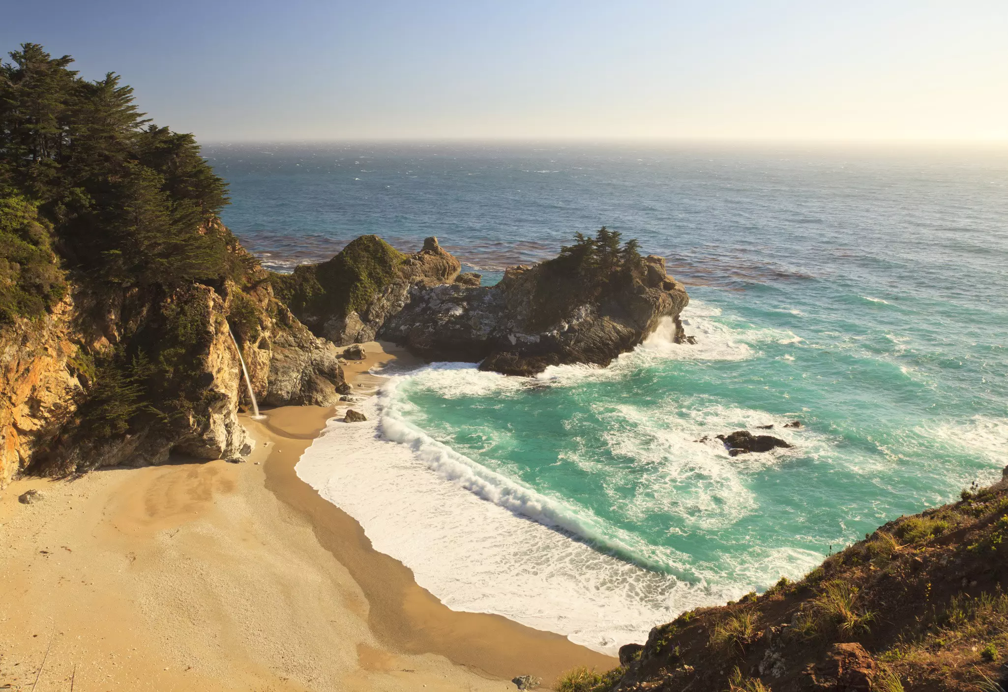 A waterfall crashes down onto a sandy cove with waves lapping at the beach.